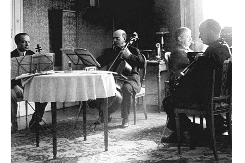 An old photograph of a group of men wearing suits performing on string instruments with sheet music in front of them.